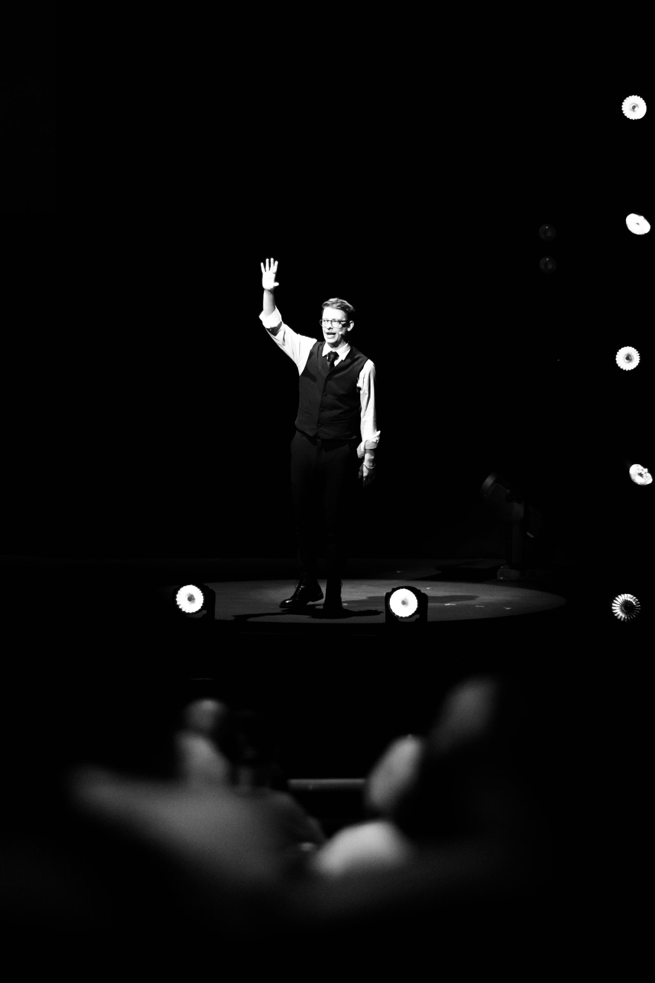 Matthew Pomeroy standing in a spotlight on a dark stage, one hand raised, addressing an audience