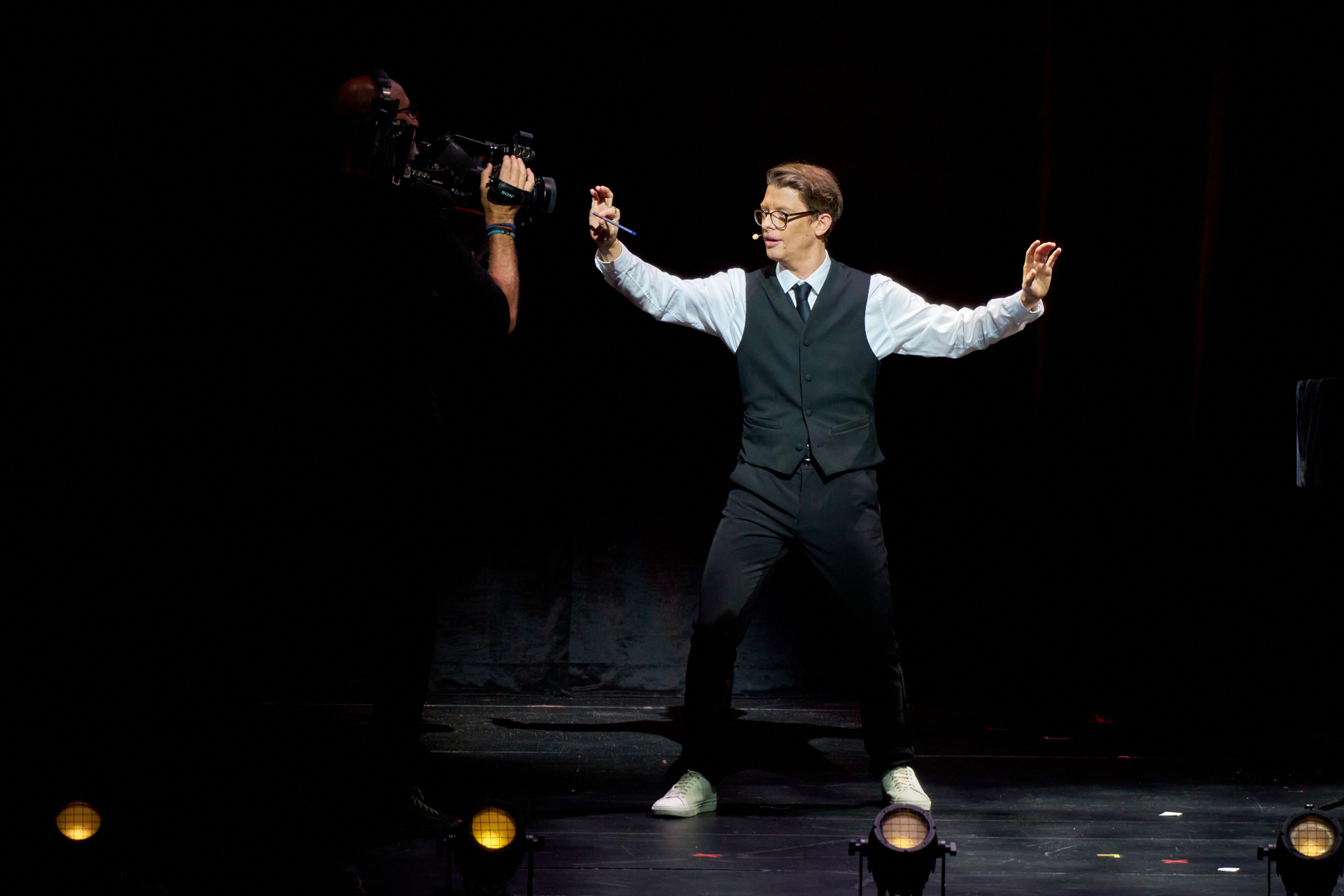 Matthew Pomeroy performing arms-out while a cameraman films on stage