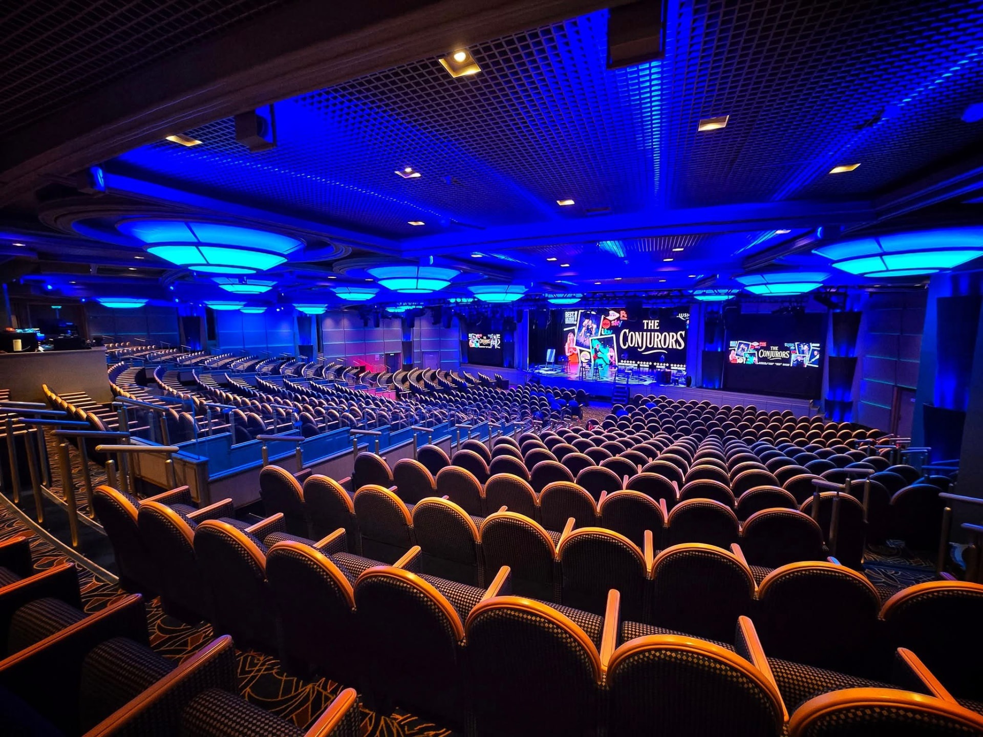 A blue-lit theatre at sea with The Conjurors on the stage screens