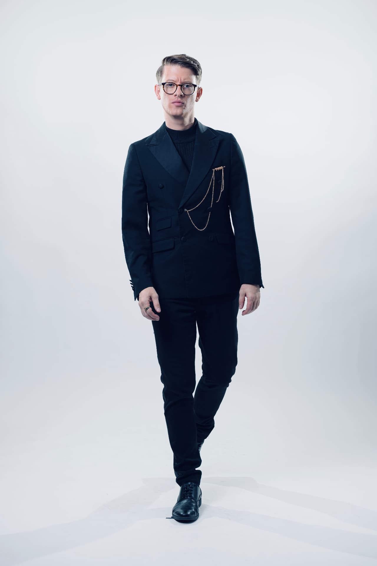 Matthew Pomeroy walking towards the camera in a black dinner jacket with gold chains, studio photograph