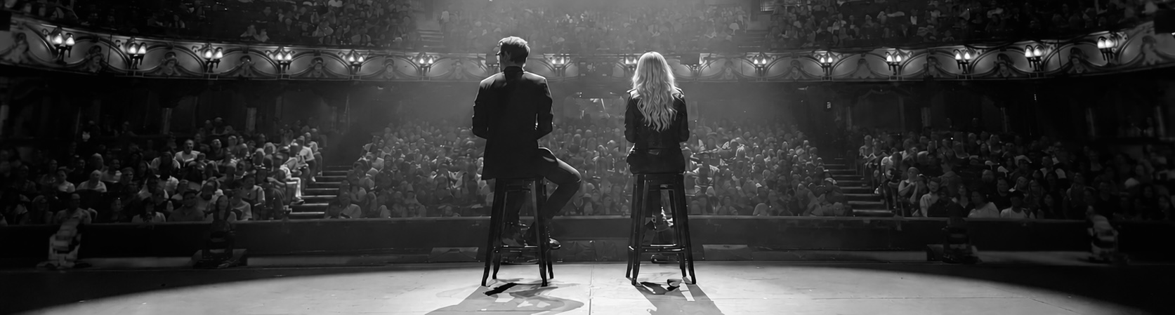 Matthew Pomeroy and Natasha Lamb seated on tall stools on the London Coliseum stage, facing a packed auditorium