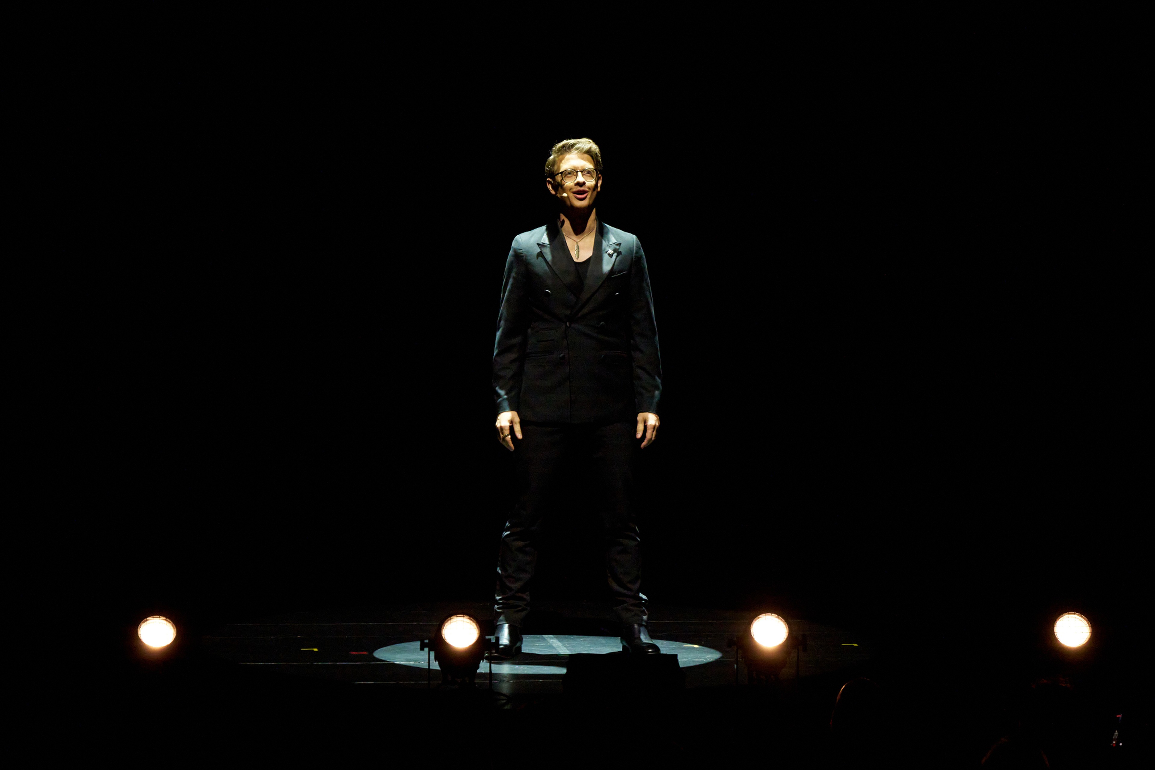 Matthew Pomeroy standing alone in a single spotlight above a row of footlights