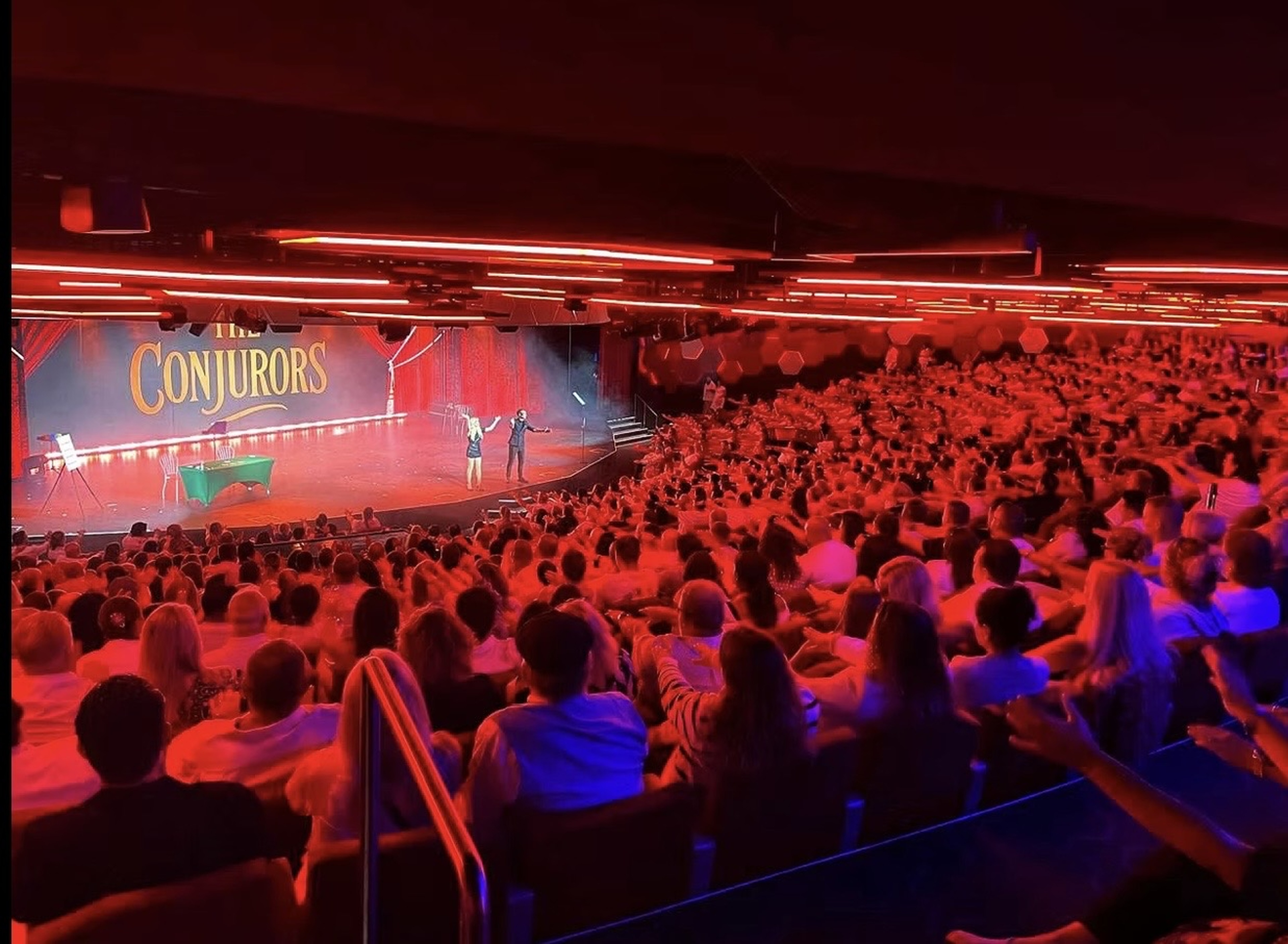 A theatre audience under red lighting facing The Conjurors on stage