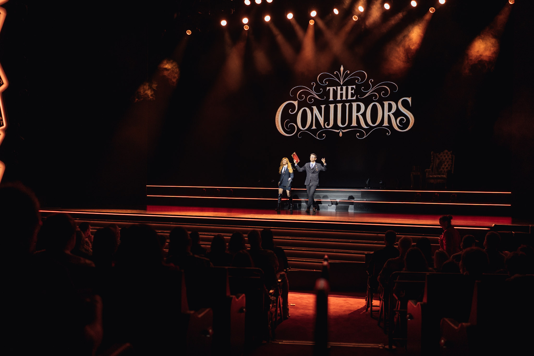 Matthew Pomeroy and Natasha Lamb greeting their audience on an orange-lit theatre stage beneath The Conjurors backdrop