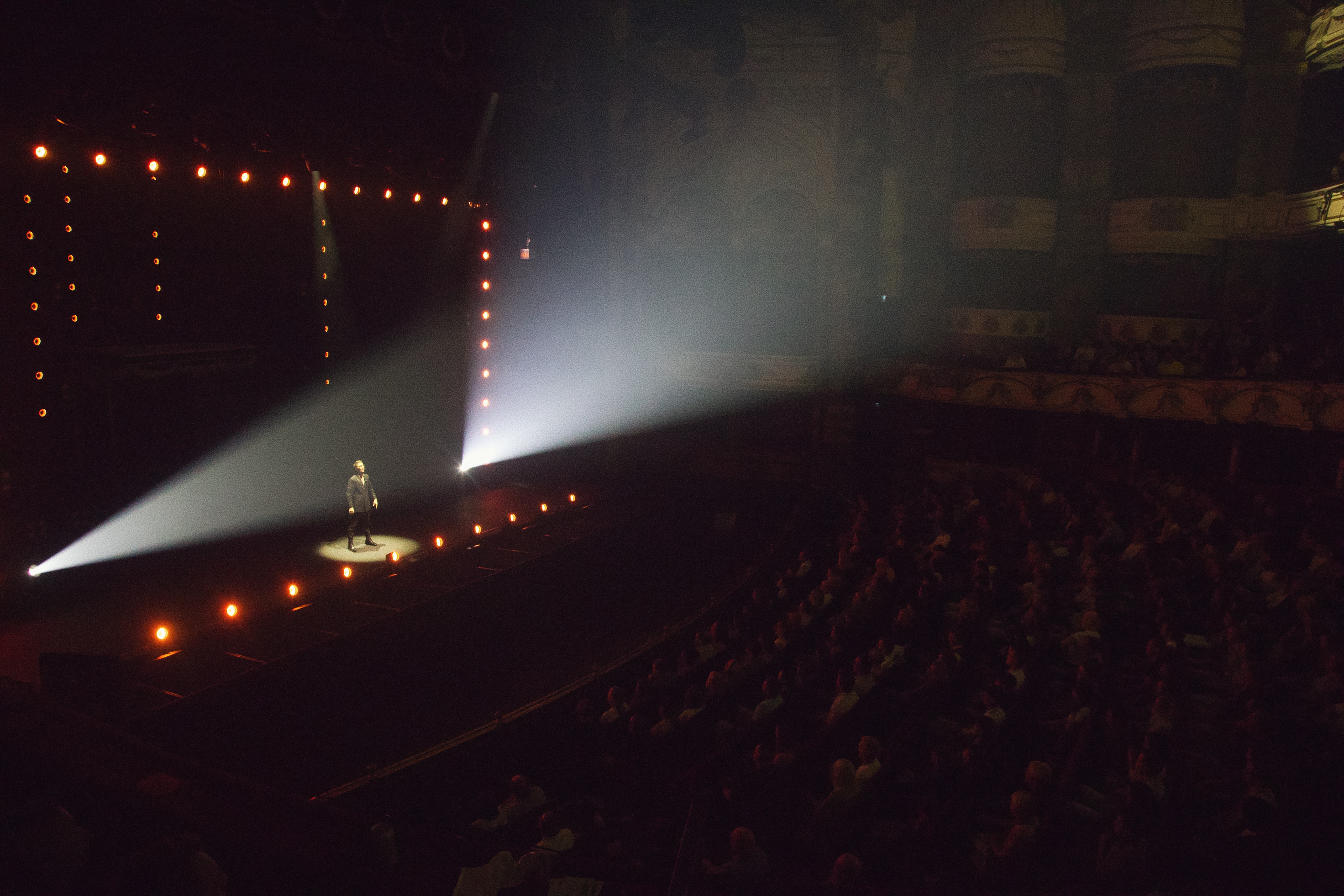 Matthew Pomeroy in a spotlight on a theatre stage, with warm footlights and a seated audience