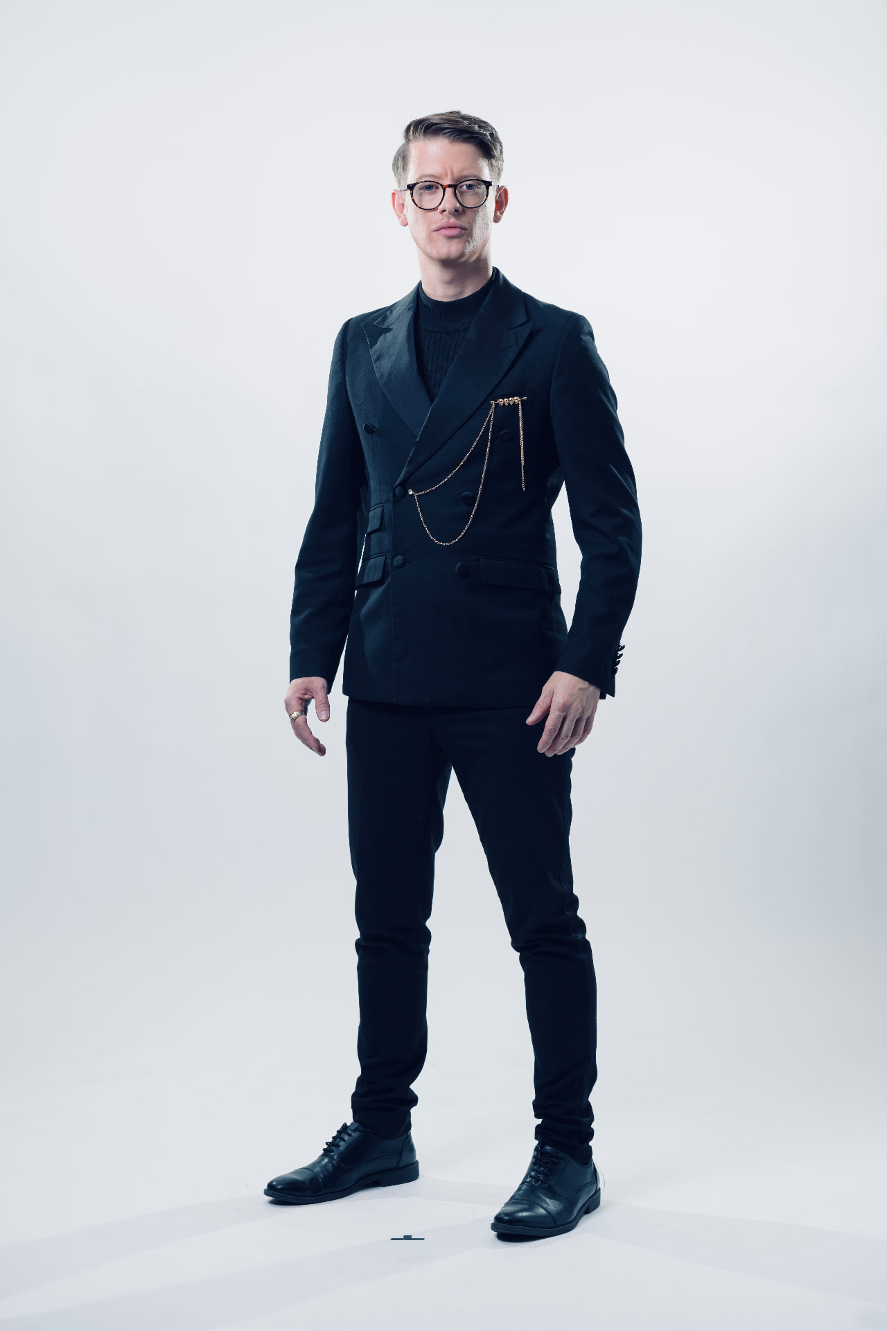 Matthew Pomeroy standing squarely in a black dinner jacket with gold chains, studio photograph